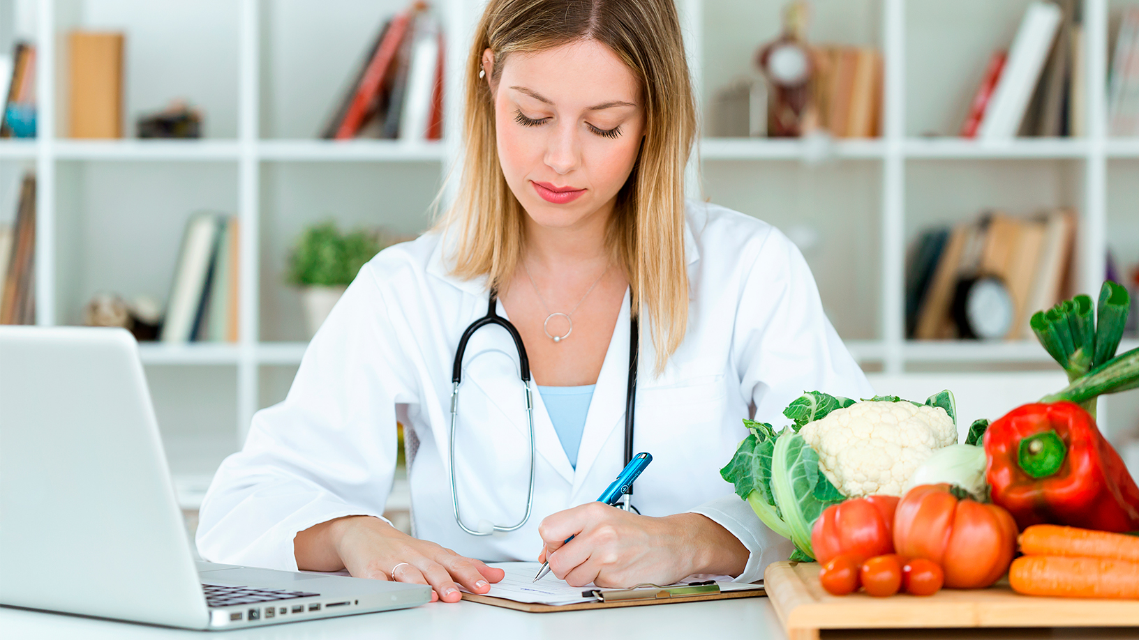 Eine Frau im weißen Kittel mit Stethoskop füllt am Schreibtisch ein Formular aus, neben sich Gemüse und ein Notebook.