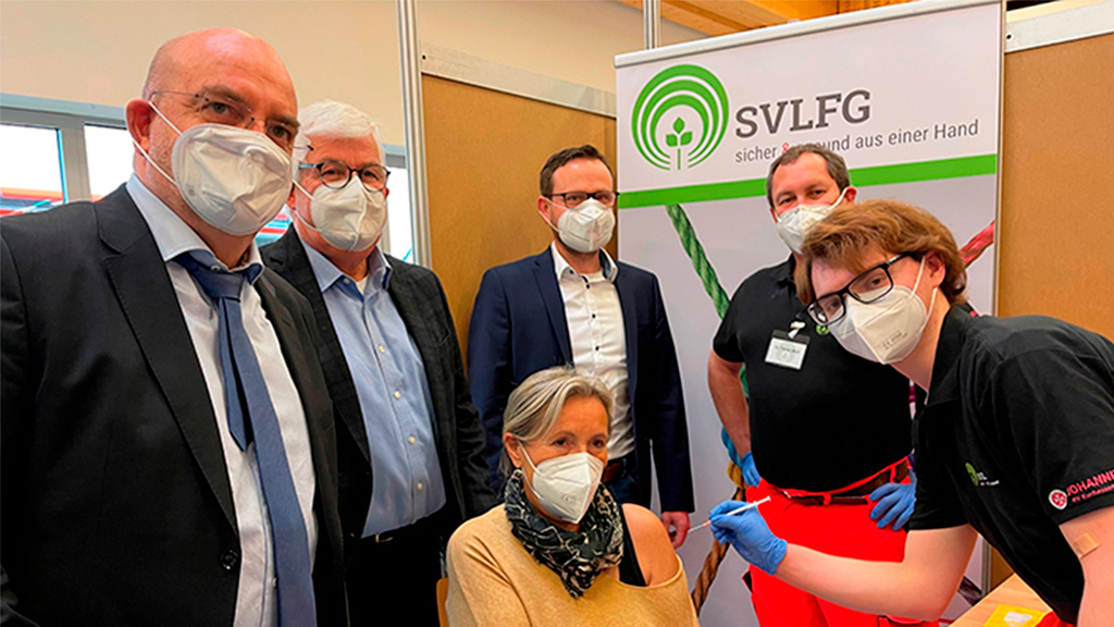 Vergrößerung des Bildes für Eine Gruppe mit Masken steht um eine Frau in der Mitte. Ein Mann hält eine Spritze an ihren Arm..