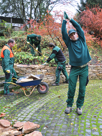 Vergrößerung des Bildes für Ein Mann hat die Arme nach oben ausgestreckt und neigt sich zur Seite, drei weitere Männer verrichten Gartenarbeit..
