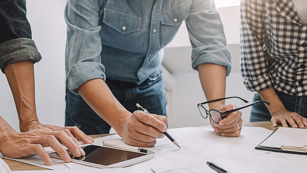 Vergrößerung des Bildes für Oberkörper und Hände von drei männliche Personen, die an einem Arbeitstisch im Gespräch sind und dabei Tablet und Unterlagen nutzen. .