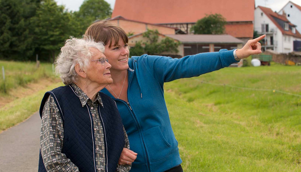 Vergrößerung des Bildes für Eine ältere Frau wird von einer jungen Frau bei einem Spaziergang in ländlicher Umgebung begleitet. Die junge Frau hält den Arm der älteren und zeigt auf etwas außerhalb des Bildes..