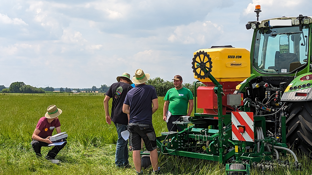 Vergrößerung des Bildes für Eine Gruppe junger Männer arbeiten auf einer Wiese. Neben der Gruppe steht ein Traktor.