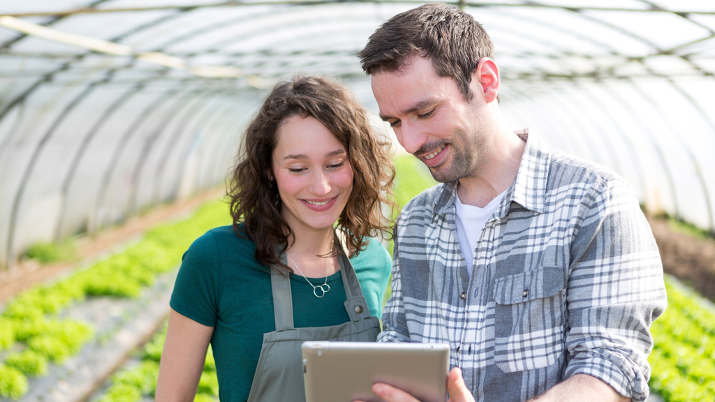 Vergrößerung des Bildes für Junger Gärtner im Gewächshaus erklärt etwas einer jungen Angestellten am Tablett..