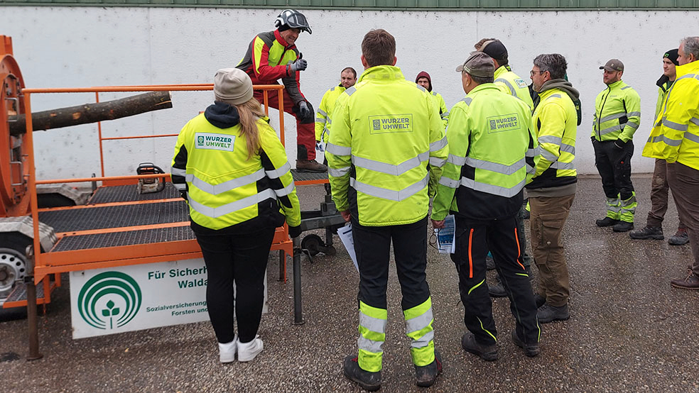 Vergrößerung des Bildes für mehrere Personen in gelben Warnjacken stehen vor einem Spannungssimulator. Eine Person in persönlicher Schutzkleidung, einschießlich Schutzhelm, steht auf dem Anhänger und erklärt..