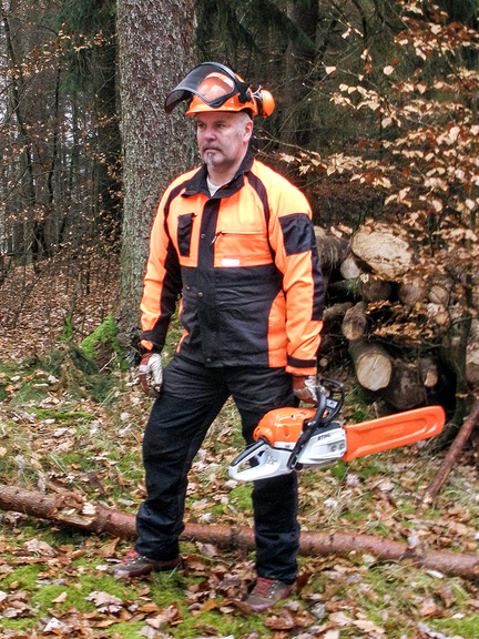 Vergrößerung des Bildes für Das Bild steht ein Mann im Wald in vollständiger Forst-PSA mit Schutzhelm mit Sicht- und Gehörschutz, Schnittschutzhose, Schnittschutzschuhen und Handschuhen. Er hält in der linken Hand eine Motorsäge..