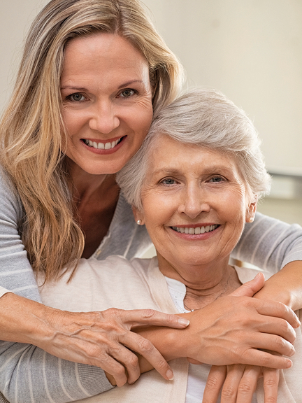 Vergrößerung des Bildes für Eine junge Frau umarmt eine Seniorin von hinten, beide strahlen in die Kamera..