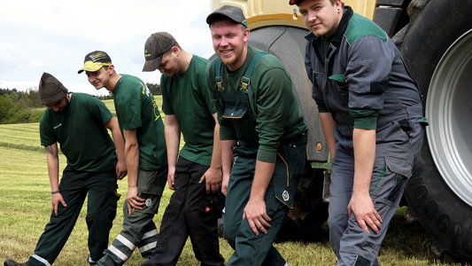 Personen dehnen im Stehen die Wadenmuskulatur. Sie stehen auf dem Feld, im Hintergrund ein Traktor