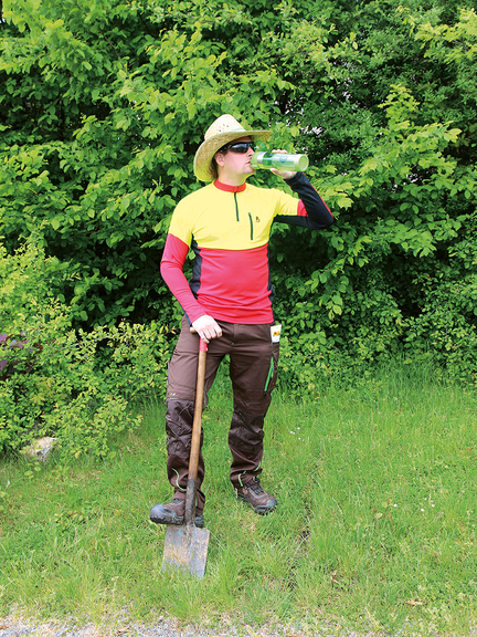 Vergrößerung des Bildes für Mann mit Strohhut und funktioneller, leichter Arbeitskleidung für den Sommer. Er hält ein Spaten und trinkt aus einer Trinkflasche. .