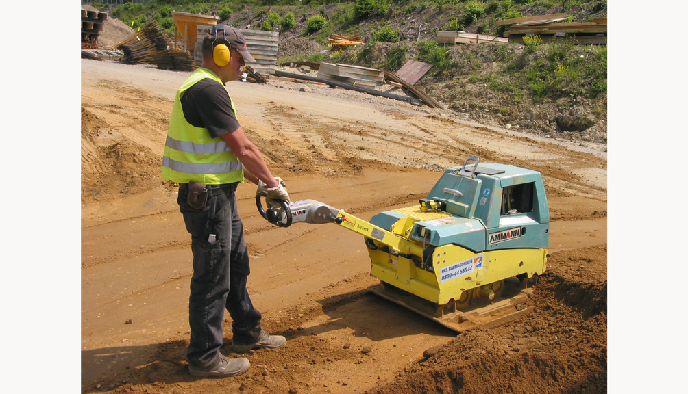 Vergrößerung des Bildes für Arbeiter mit Körperschutzkleidung verdichtet Boden mit Rüttelplatte.