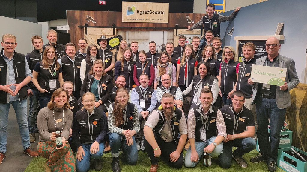 Vergrößerung des Bildes für Gruppenfoto junger freundlicher Menschen mit einheitlicher Weste und Namenschildern auf den Westen auf Messestand. An der hinteren Wand ein Schild mit der Aufschrift. Agrar-Scouts. Ein Mann im Anzug hält ein Schild: SafeHabitus..