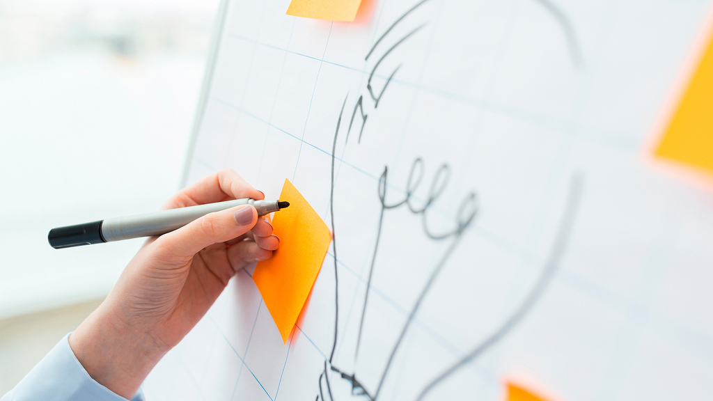 Vergrößerung des Bildes für Flipchart mit der Skizze einer Glühbirne. Darüber klebt ein orangener Klebezettel, der von einer Person beschriftet wird. .