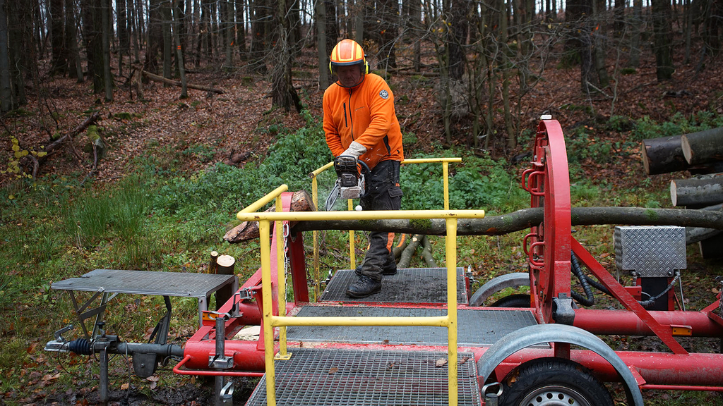 Vergrößerung des Bildes für Mann in persönlicher Schutzausrüstung steht auf Spannungssimulator. Er zersägt einen Baumstamm mit der Motorsäge..